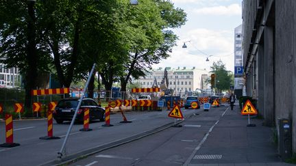 Tvåfilig väg där ena körfältet är avstängt. Byggstängsel på ena sidan av vägen och flera vägmärken som leder om trafiken och varnar för vägarbeten och vakt längre fram.