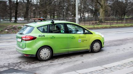Bild på en grön Färdtjänstbil i storlek med en personbil. Den är ute och kör på vägen.
