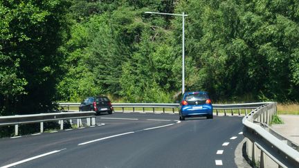 En svart och en blå bil kör bort från tittaren på Bergsjövägen. Det är en solig dag och grön skog vid sidan av vägen.