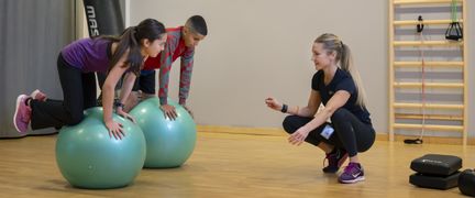 Två unga personer balanserar på balansboll. En instruktör sitter på huk bredvid.