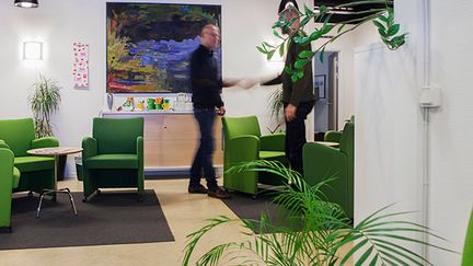 fyra gröna fåtöljer i ett väntrum. I bakgrunden står två män och prata med varandra.