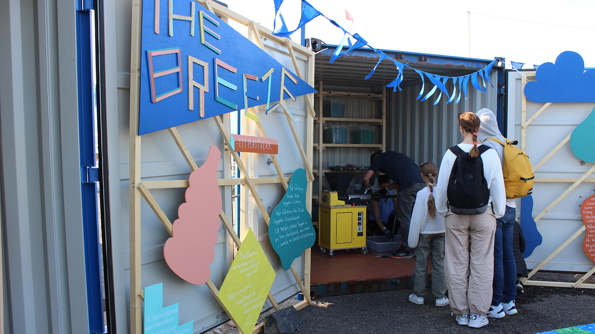 Två vuxna och ett barn kikar in i en öppen container med blåa vimplar och färgglada skyltar. Inuti står en människa och jobbar med en gul maskin.