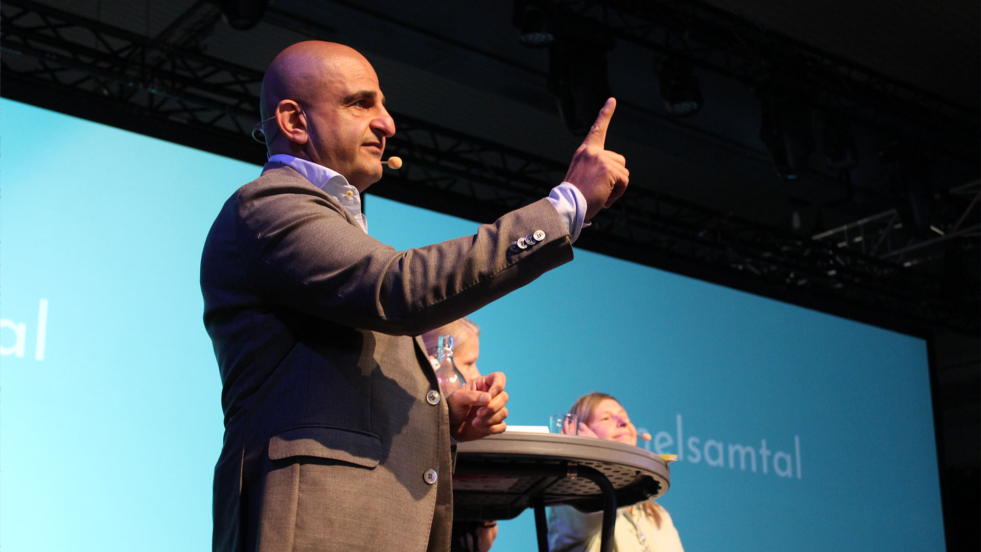 En person håller upp ett finger i luften samtidigt som han pratar framför stora skärmar på scen.