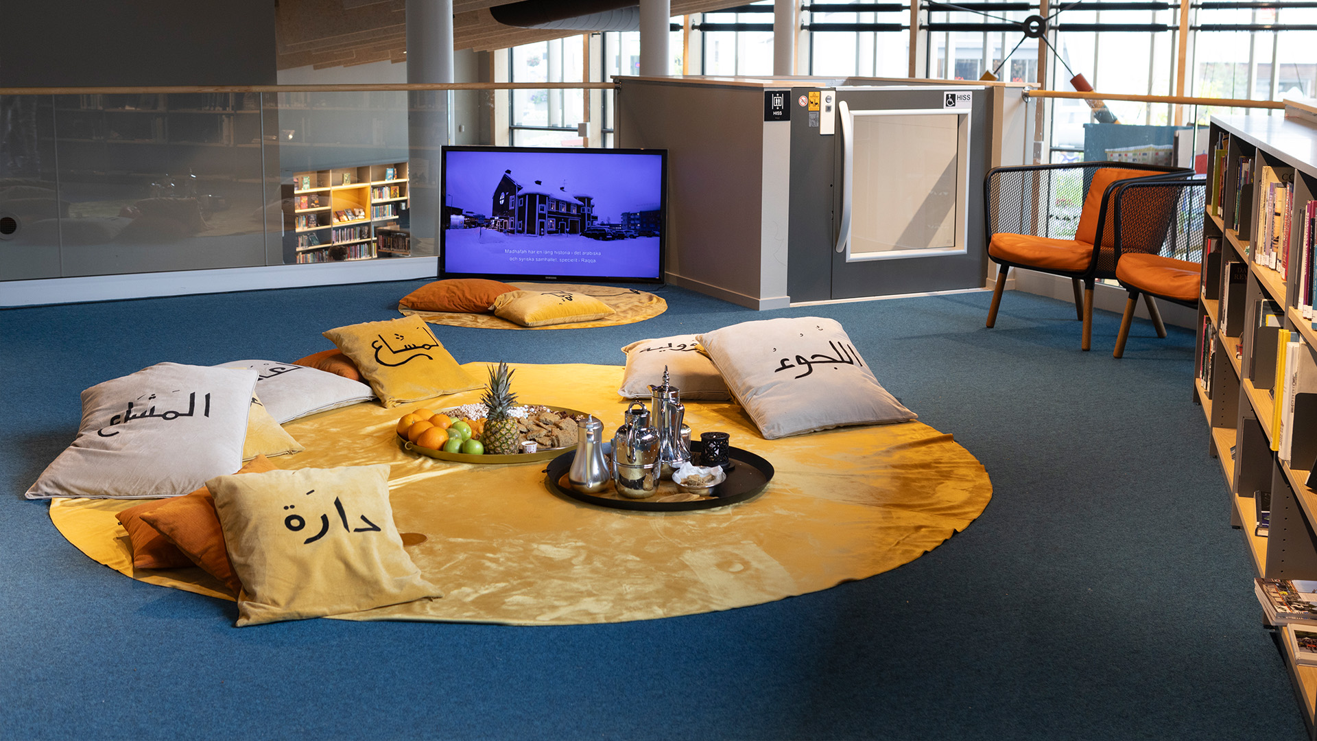En rund gul matta ligger på ett golv bredvid en bokhylla, på den finns kuddar med arabiska tecken, en teservis och ett frukfat.