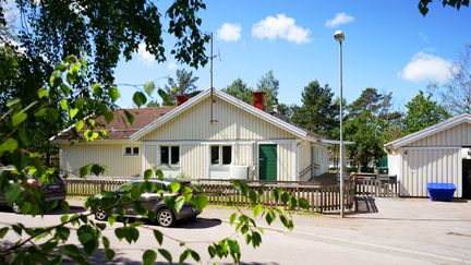 Ett träd i förgrunden och en vitt trähus i bakrunden. Det är sommar.