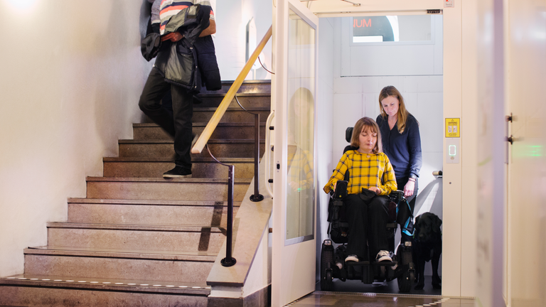 En kvinna i rullstol kommer ut ur en hiss samtidigt som en annan person går ner för trappan bredvid hissen.
