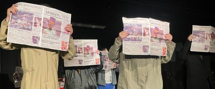En teaterföreställning där fem personer håller upp en tidning framför sig.