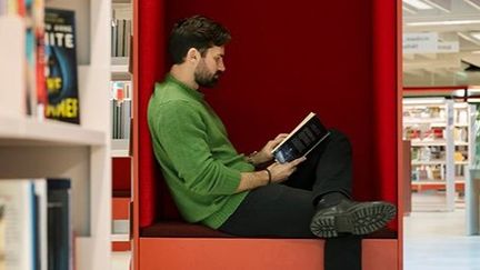 En man sitter på biblioteket och läser en bok i en röd biblioteksmöbel.
