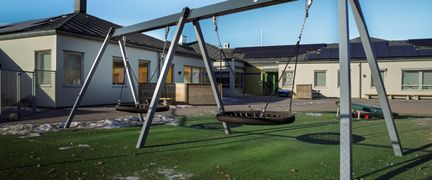 En lekplats med några gungor framför Lilla Trulsegårdsskolan. I bakgrunden kan några skolbyggnader ses.