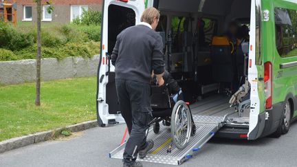 En man kör en person i rullstol mot en grön färdtjänstbuss som har en utrullad ramp.