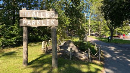 En träskylt med texten Hjällbo parklek. I bakgrunden ser du en lekplats omgiven av gröna träd och och grönt sommargräs.
