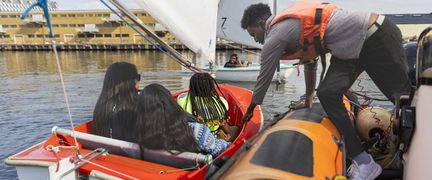 En kille står på en brygga och håller i en röd jolle. I jollen sitter tre tjejer. 