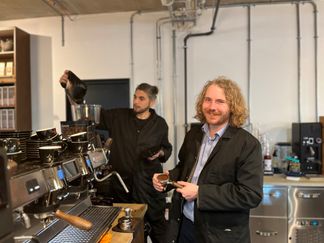 en man fyller på kaffebönor i en maskin och en håller en kaffepuck och ler mot kameran