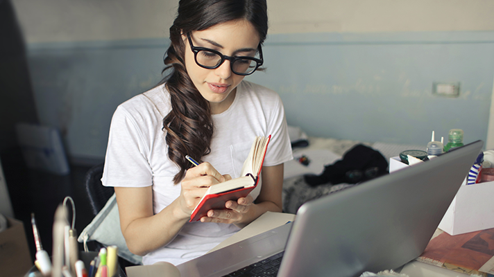 En kvinna sitter vid ett skrivbord och antecknar i en anteckningsbok. Det står en dator på skrivbordet.