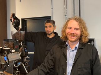 två män står vid kaffemaskin och ler mot kameran