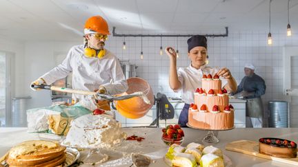 Ett kaosartat konditorikök där en man står och skyfflar mjöl i en cementblandare och kvinnan bredvid hon försöker dekorera en tårta.