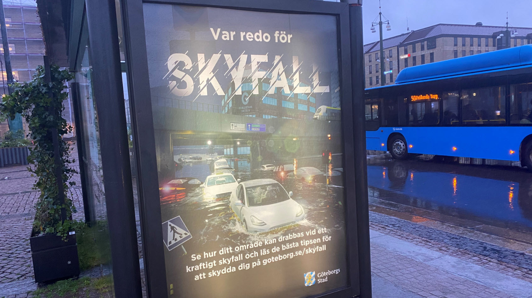Informationstavla vid en regnig busshållplats som visar affisch med en visualisering av ett översvämmat Gamlestaden