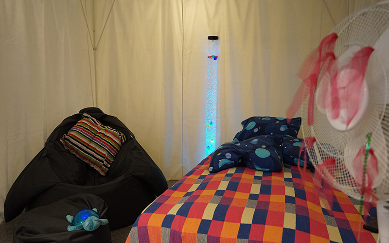 Ett rum med säng som har rutigt överkast, en svart saccosäck och en fläkt med rosa band i. I bakgrunden står ett vattenrör.