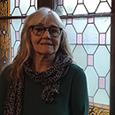 Eva Johansson ler in i kameran, bakgrunden är ett fönster med färgade glasrutor i olika former