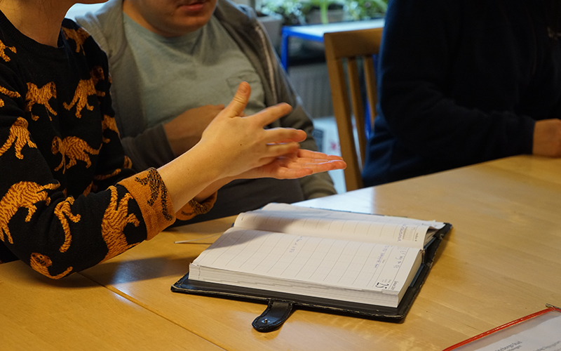 En person kommunicerar med teckenspråk till två andra personer. På bordet framför ligger en kalender.