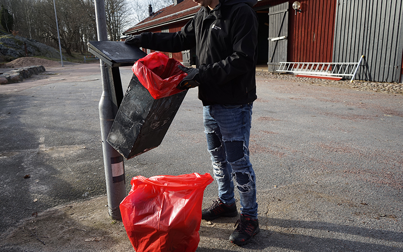 En person står utomhus och tömmer en papperskorg som hänger på en lyktstolpe.
