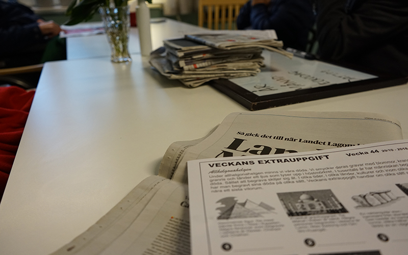 Personer sitter kring ett bord med tidningar på. Det ligger ett papper på bordet som det står "veckans extrauppgift" på.