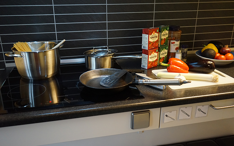 En köksbänk med mat på, förpackningar med krossade tomater och olika grönsaker. En spis med en stekpanna och en kastrull med spagetti i.