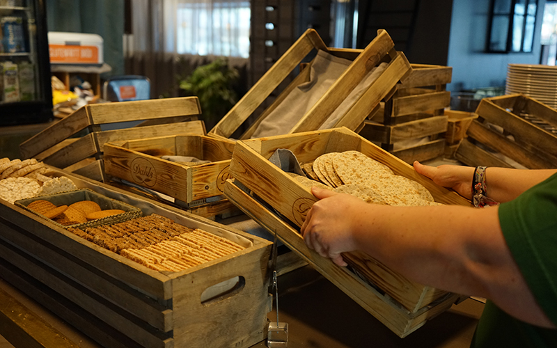 En person lyfter en brödkorg med knäckebröd i. På ett bord står flera korgar med olika sorters bröd i.